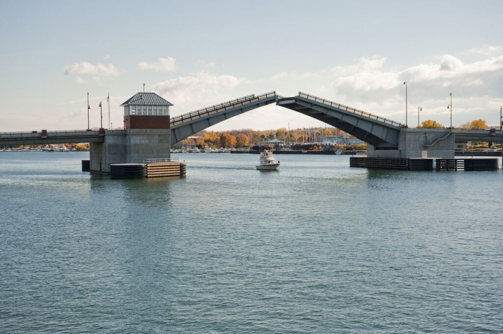 A boat passes under a raised drawbridge over a calm body of water with a small building on the bridge and autumn trees in the background.