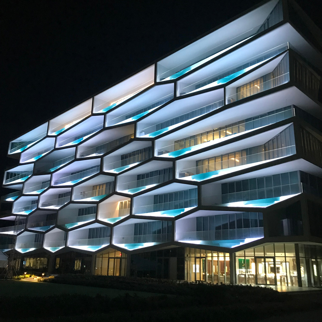 Modern multi-story building at night with illuminated hexagonal balconies and large windows.