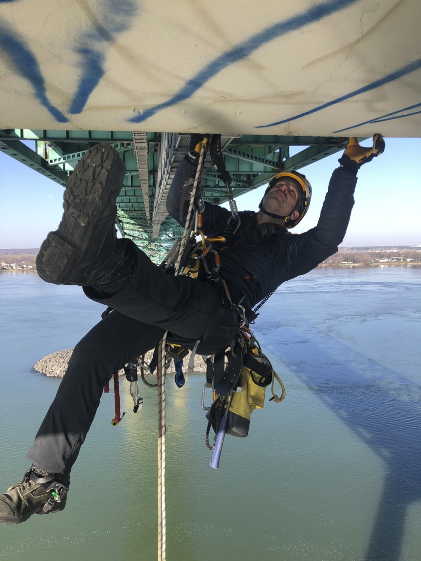 A worker in safety gear performs maintenance under a bridge, suspended by ropes over a body of water.