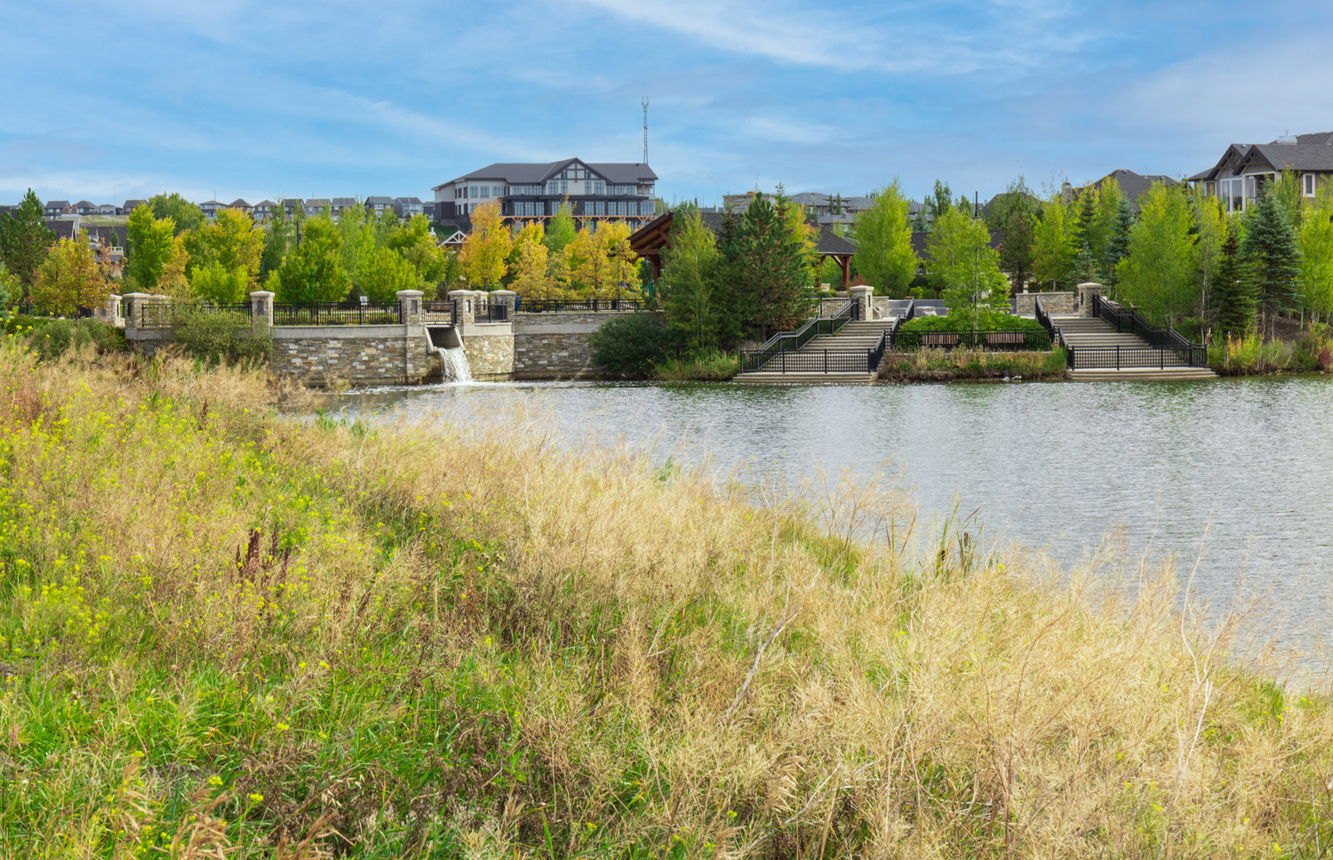 Park scene with a pond, grassy foreground, and a stone bridge with steps leading to a gazebo. Trees and residential buildings are in the background under a blue sky.