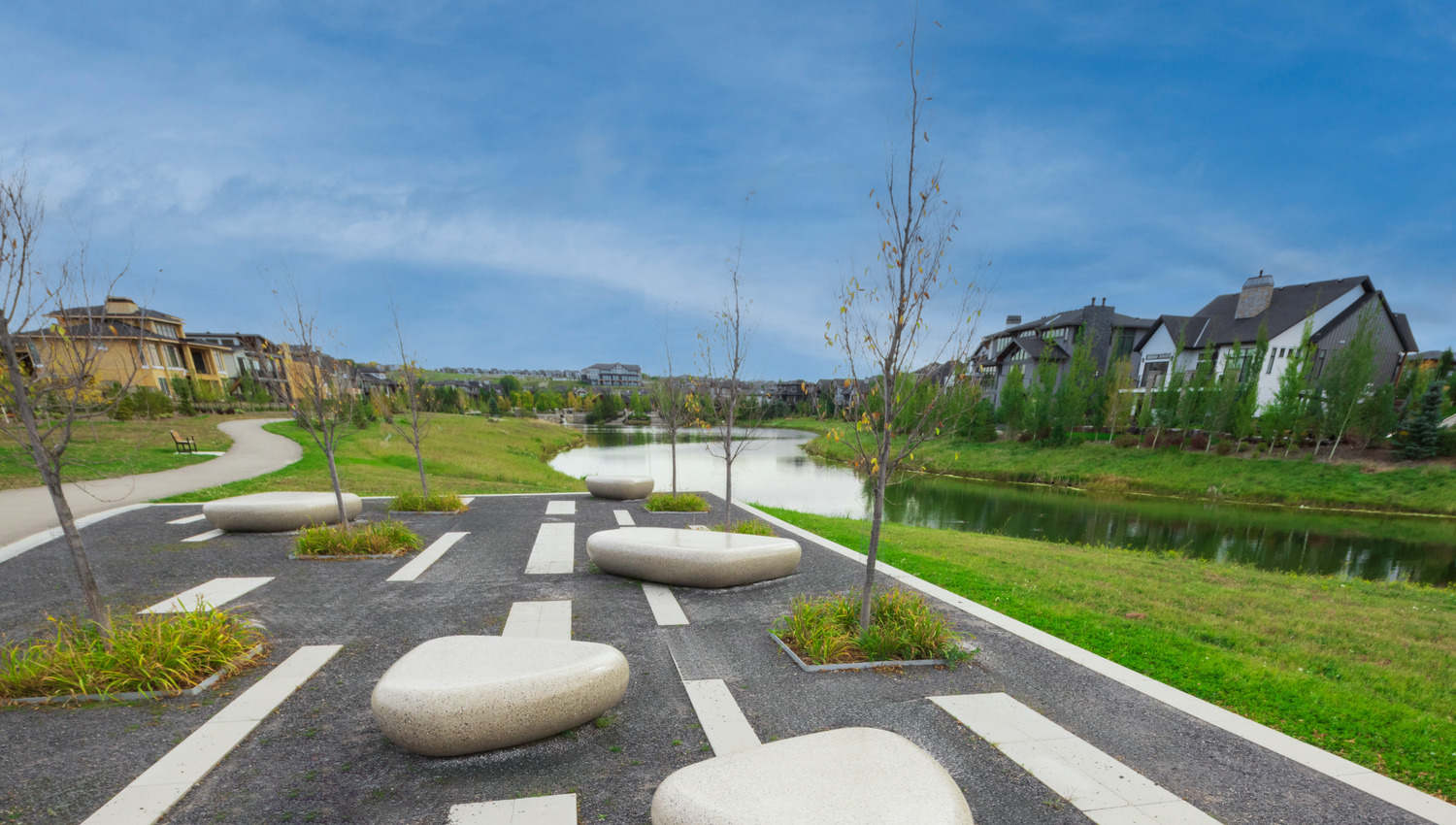 A modern, landscaped park with stone seating, young trees, and a winding path by a tranquil pond, surrounded by houses under a blue sky.