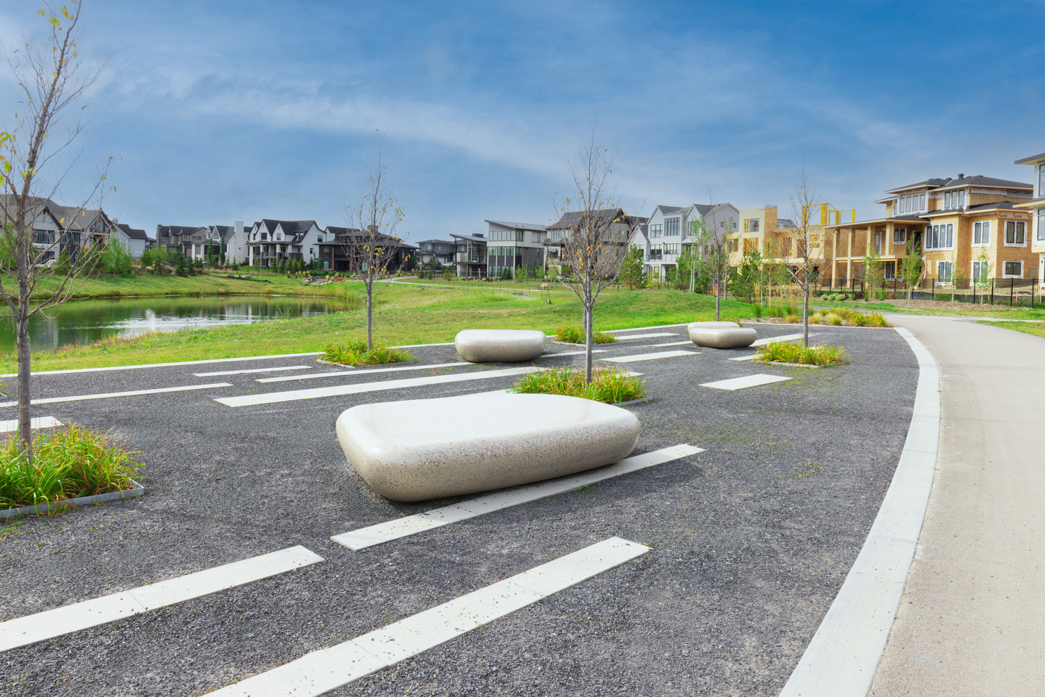 A modern outdoor area with large stone benches, young trees, and a path. Nearby are contemporary residential buildings with a pond in the background under a clear blue sky.