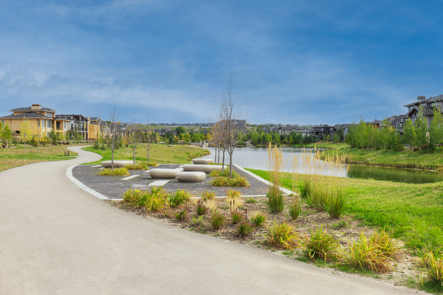 A paved path curves through a landscaped park with grass, small trees, and shrubs beside a pond, under a partly cloudy sky.