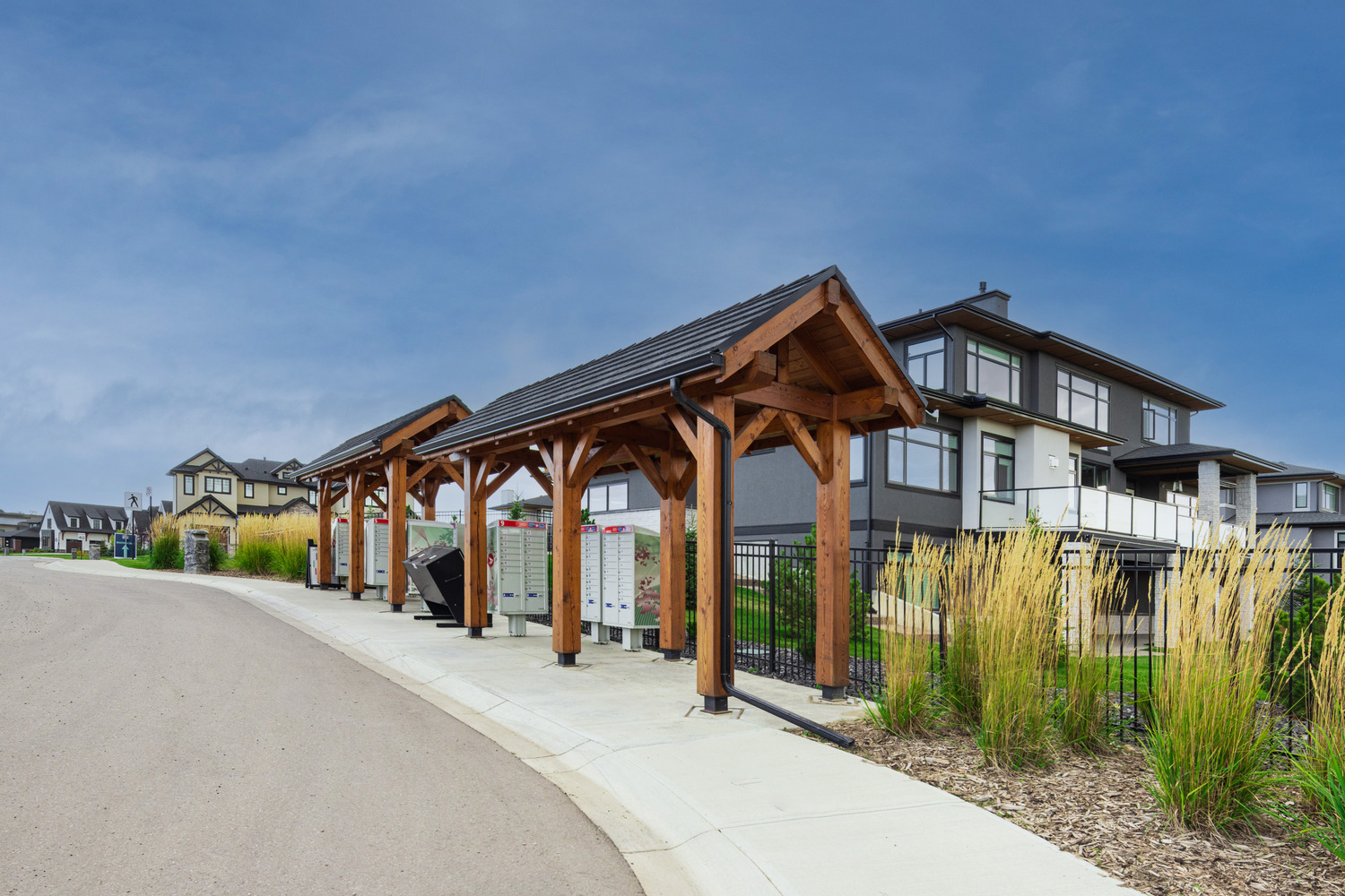 Residential street with a pavilion and mailboxes, next to modern houses and landscaped greenery under a blue sky.