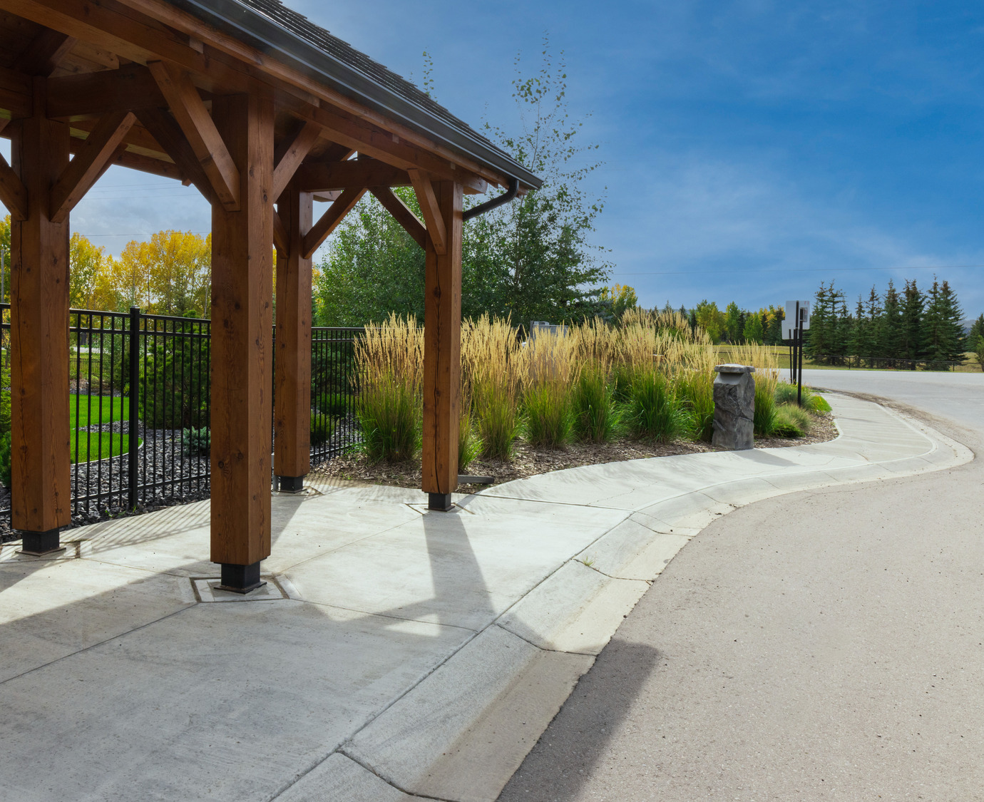 Covered wooden structure next to a road with landscaping, including tall grasses and trees in the background.