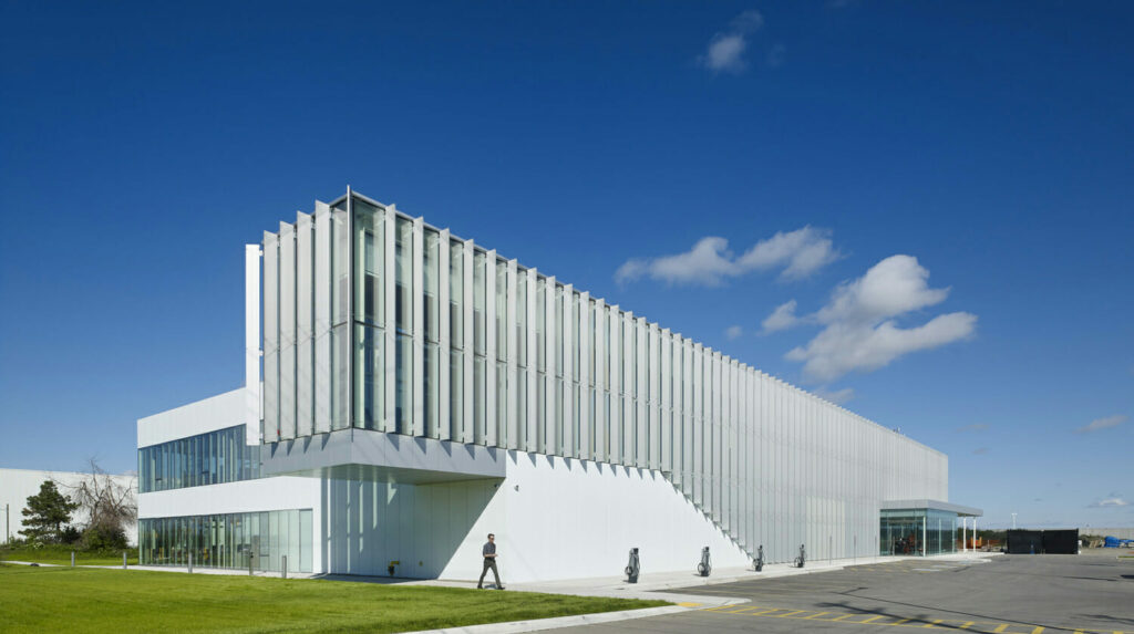 Un bâtiment blanc moderne avec des lattes verticales, de grandes fenêtres en verre et un escalier extérieur se dresse sous un ciel bleu clair. Une personne marche dehors sur une zone pavée.