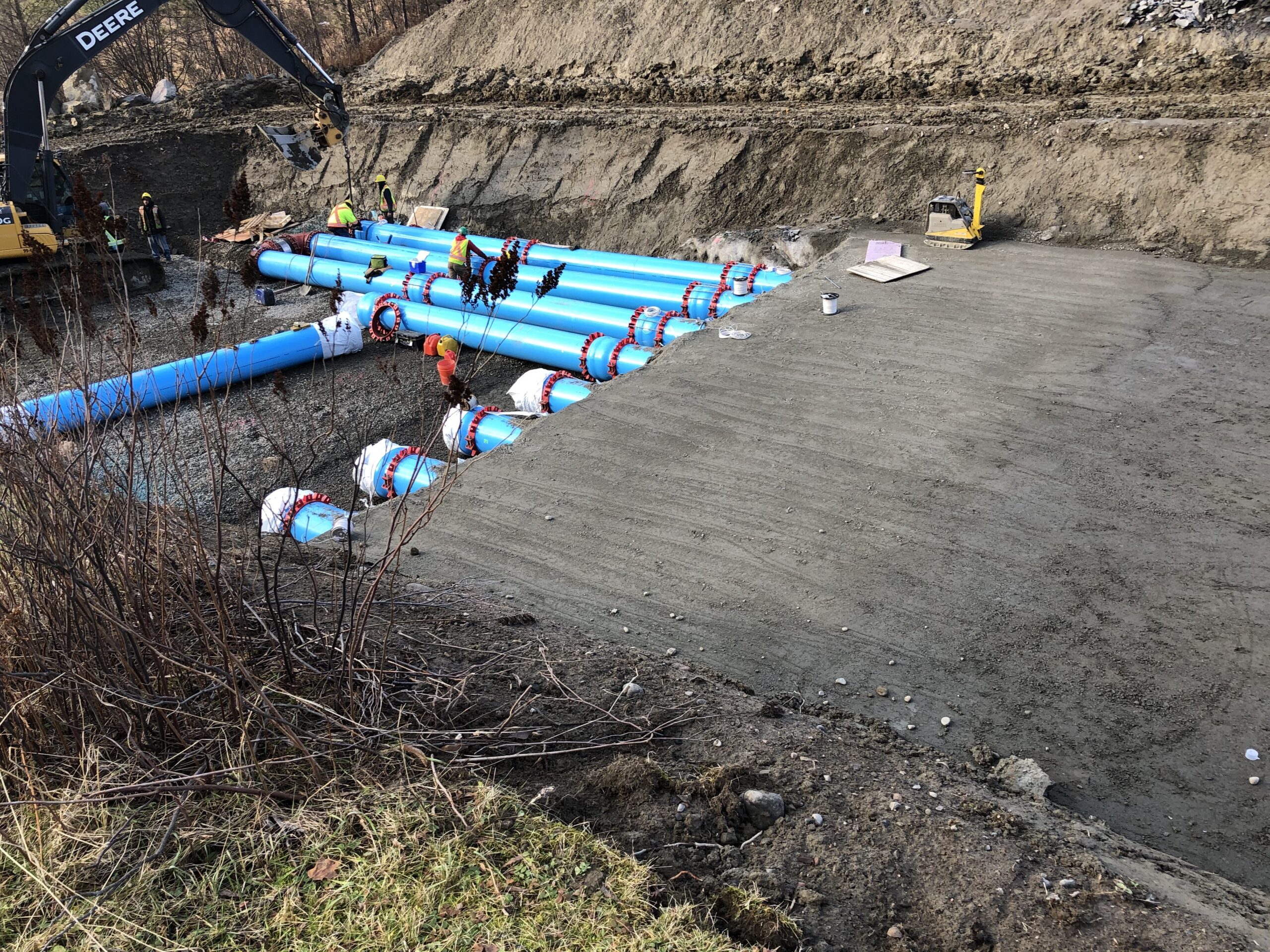 Construction site with workers installing large blue pipes in a trench. An excavator and construction materials are visible nearby.