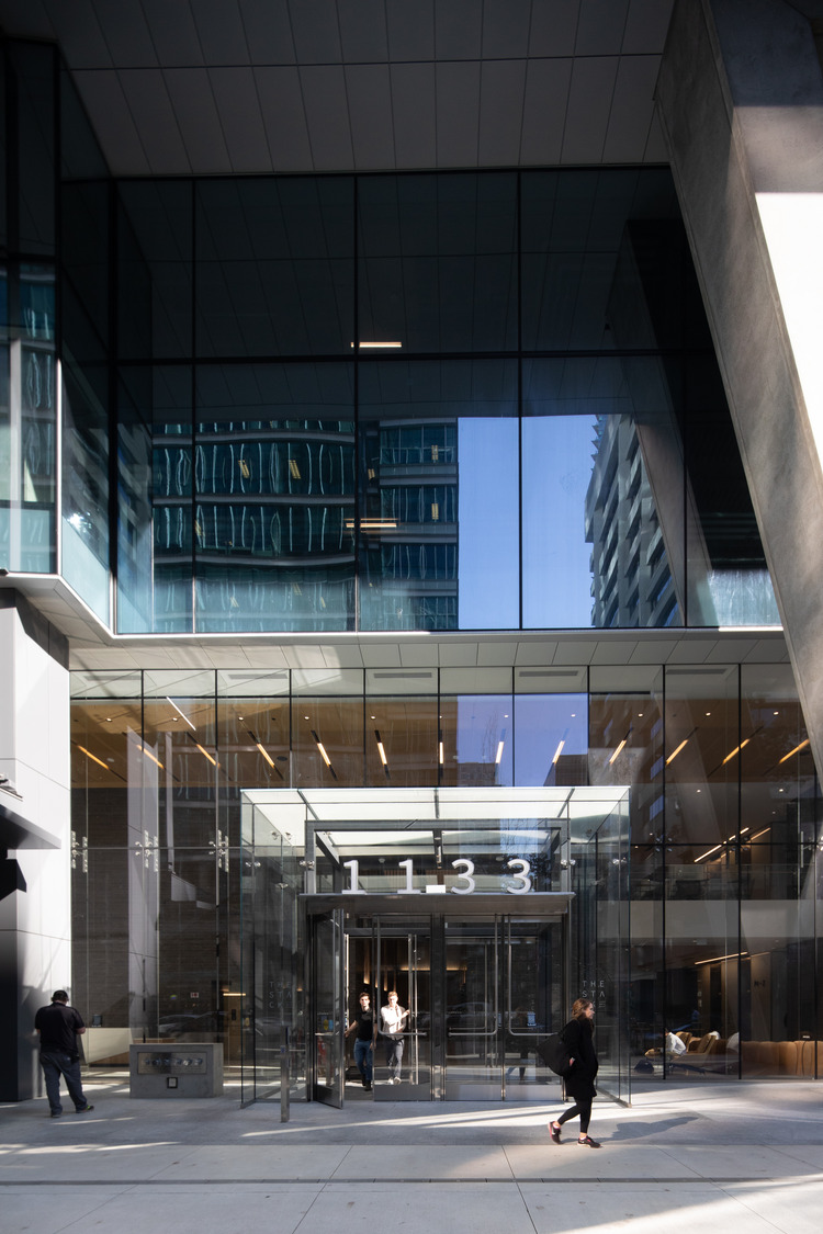 Entrée d'un bâtiment moderne en verre avec le numéro « 1133 » au-dessus de la porte. Les piétons marchent à l'extérieur du bâtiment par une journée ensoleillée. Les reflets des bâtiments voisins sont visibles dans la façade vitrée.