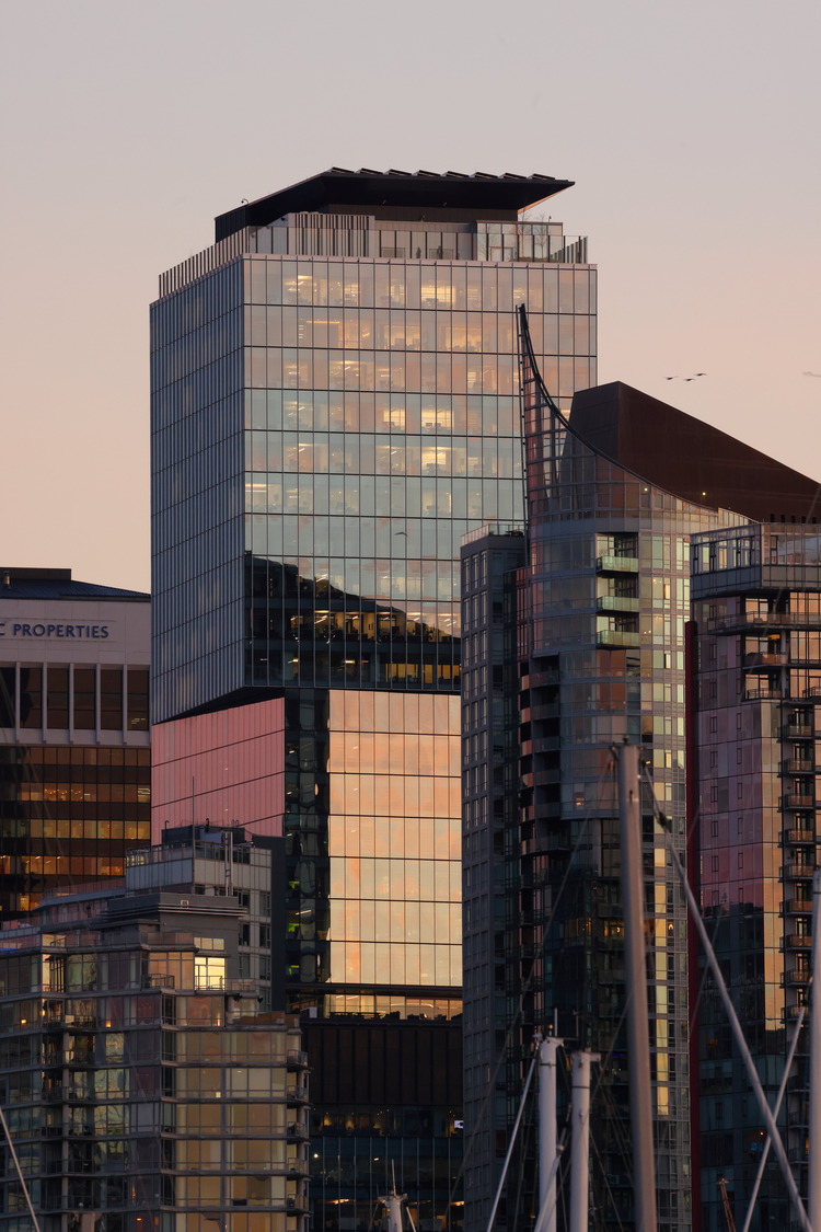 De grands bâtiments modernes aux façades de verre réfléchissantes se dressent sur un ciel clair au coucher du soleil, capturant des teintes roses et oranges.