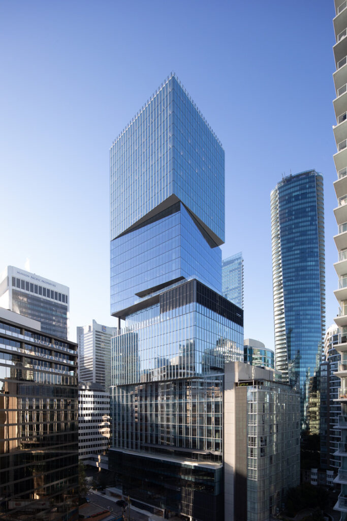 Un gratte-ciel moderne en verre aux sections décalées se dresse parmi d'autres immeubles de grande hauteur sous un ciel bleu clair dans un environnement urbain.