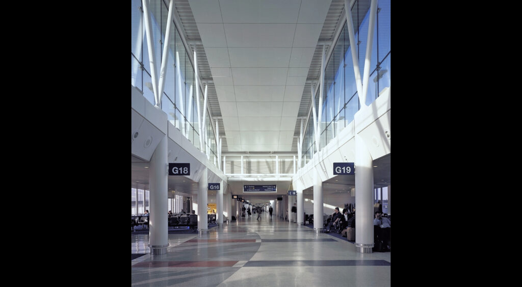 Long couloir du terminal de l'aéroport avec de hauts plafonds et de grandes fenêtres. Les portes G18 et G19 sont visibles, ainsi que des rangées de sièges et des passagers dispersés attendant leur vol.