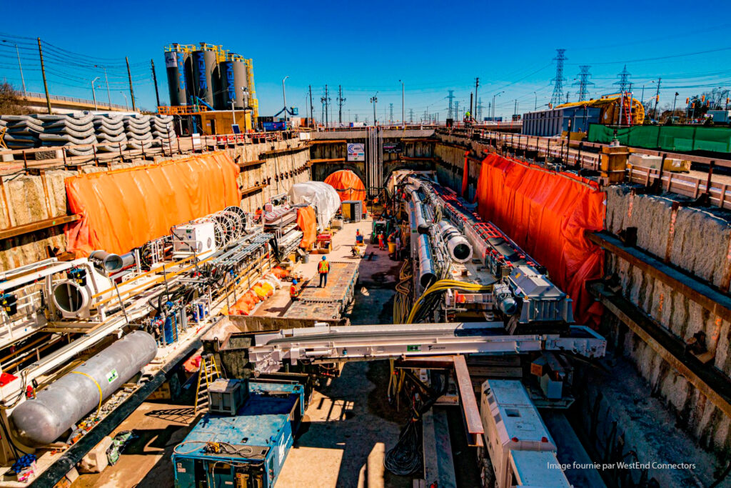 Chantier de construction avec machines et ouvriers construisant un tunnel souterrain. Des bâches orange sont drapées sur les côtés et divers équipements lourds sont visibles. Les lignes électriques et les réservoirs de stockage sont en arrière-plan.