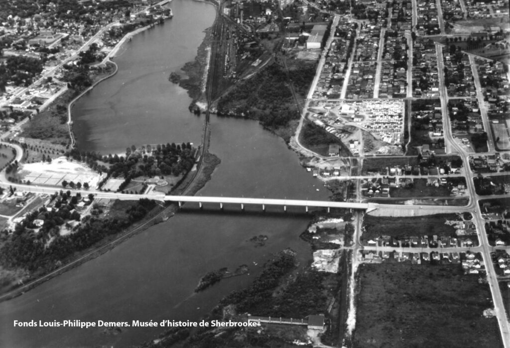 Vue aérienne d’une ville traversée par une rivière. Un pont traverse la rivière, reliant deux parties de la ville. Des maisons et des bâtiments sont visibles des deux côtés de la rivière.