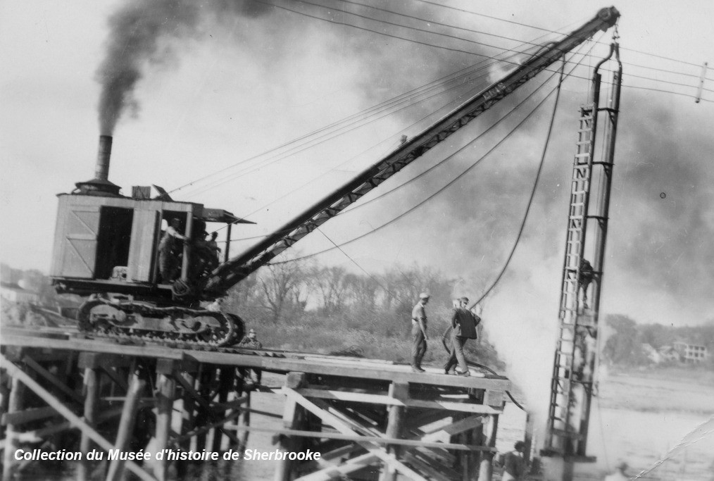 A crane operates on a wooden bridge under construction, with two workers nearby and smoke emitting from the machinery. Image credited to Collection du Musée d'histoire de Sherbrooke.