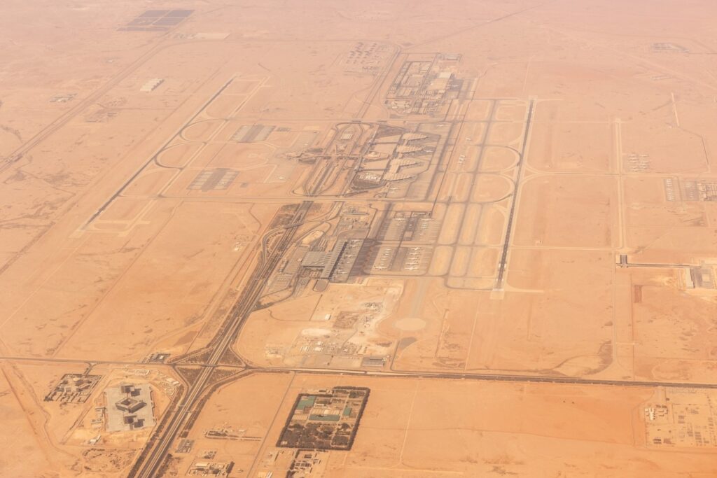 Aerial view of a large, modern airport surrounded by desert. Multiple runways, terminals, and structures are visible, as well as surrounding roads and buildings.