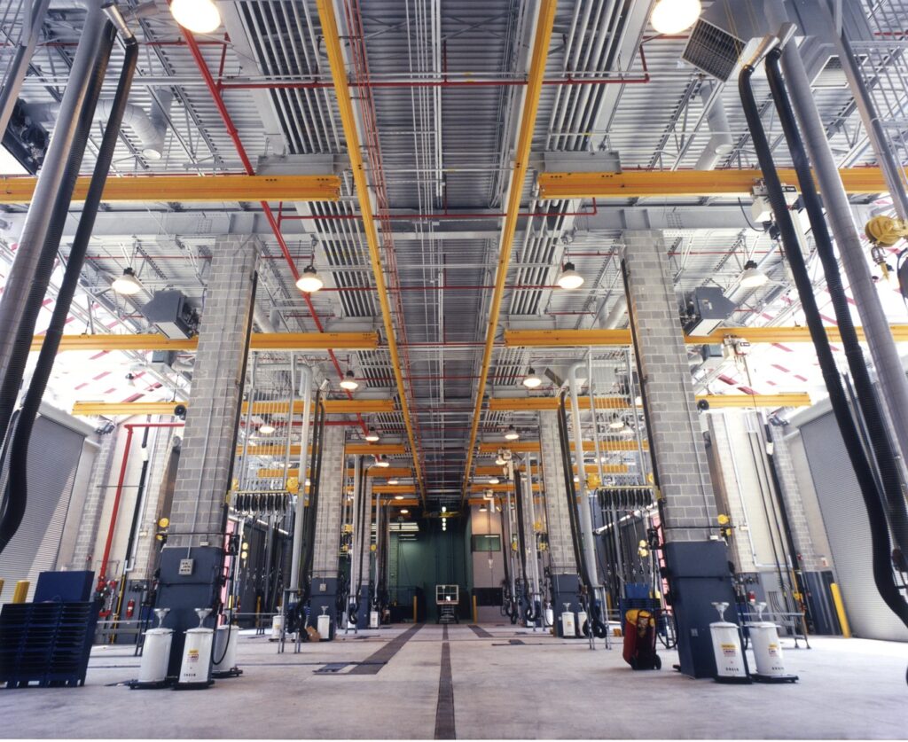 Interior of a modern industrial bus maintenance facility with high ceilings, equipped with multiple service bays and mechanical tools.