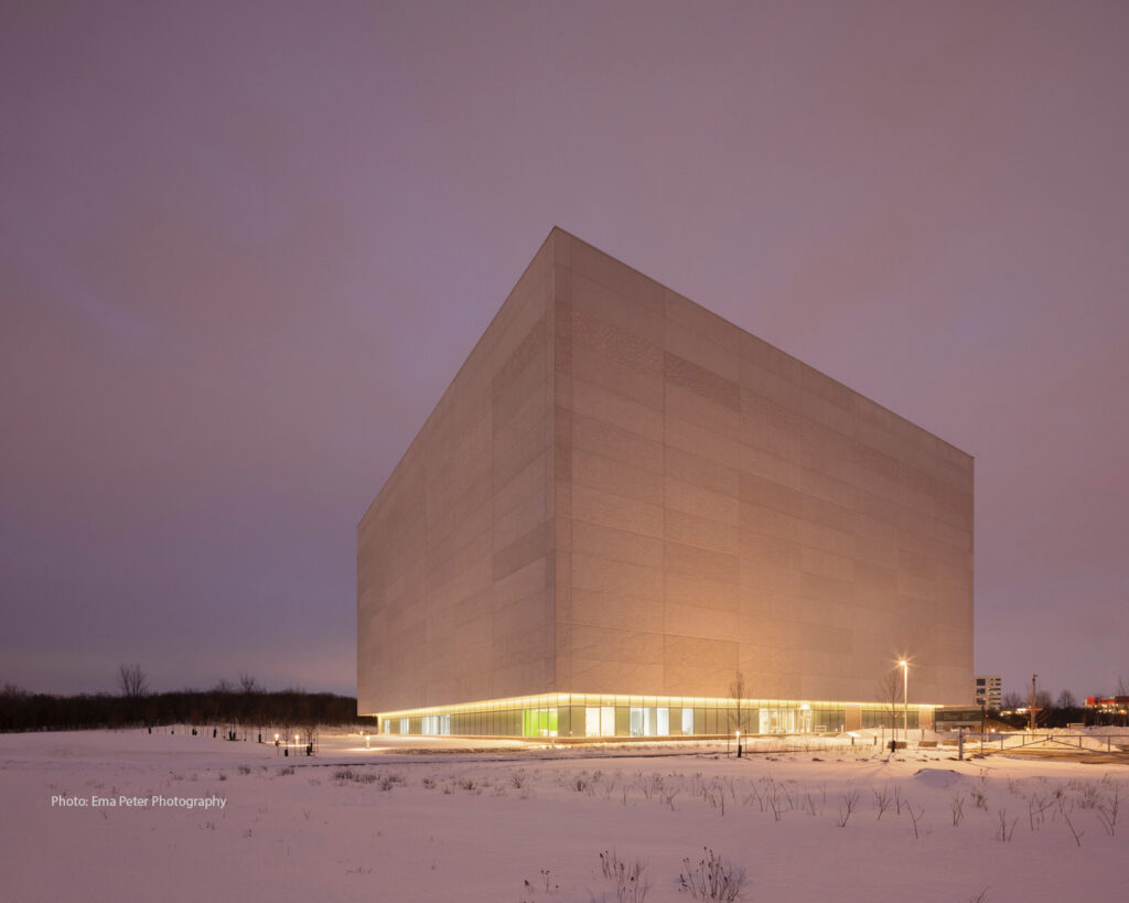 Un grand bâtiment rectangulaire illuminé se dresse sur un paysage enneigé au crépuscule, avec un ciel nuageux au-dessus. Des plantes et des lampes clairsemées sont visibles autour de la structure.