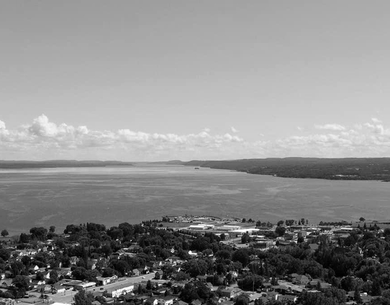 Vue aérienne en noir et blanc d'une ville au bord d'un lac avec des zones résidentielles et des arbres, s'étendant vers un grand lac sous un ciel nuageux.