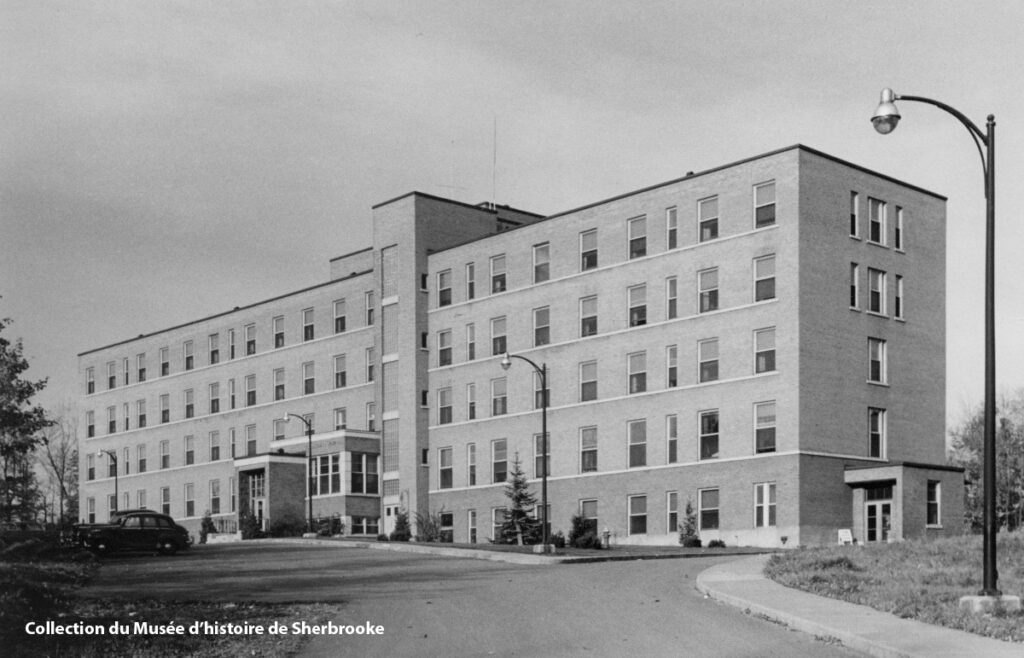 Black and white photograph of a rectangular four-story brick building with multiple windows, a main entrance with steps, and street lamps in front. Collection du Musée d’histoire de Sherbrooke.