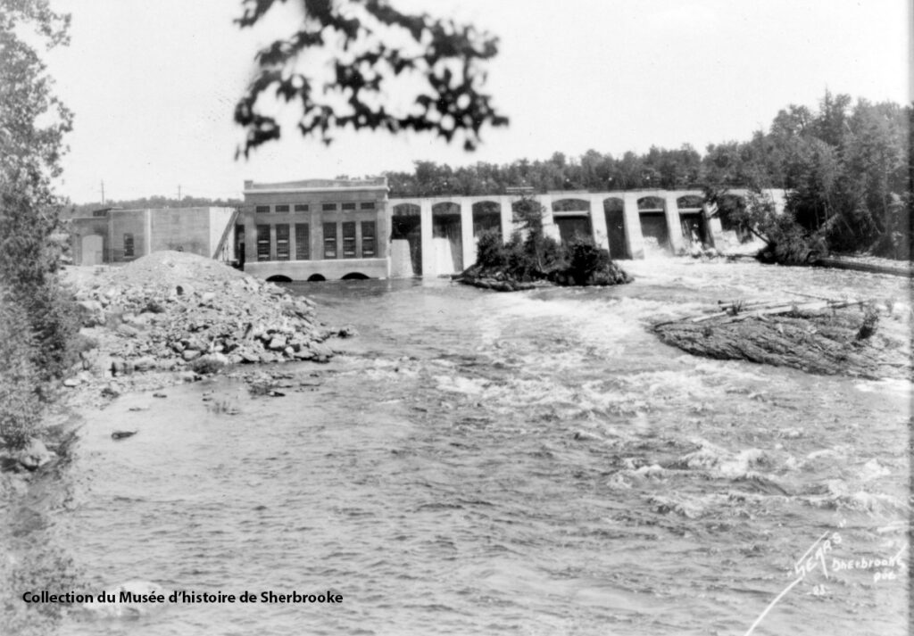 Black and white photo of the Hydro-Sherbrooke dam with water flowing over and around it, surrounded by trees. Text in the lower left corner mentions the collection of the Musée d'histoire de Sherbrooke.