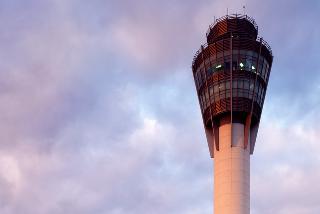 A large airport control tower stands tall against a cloudy sky, illuminated by exterior lights at dusk.