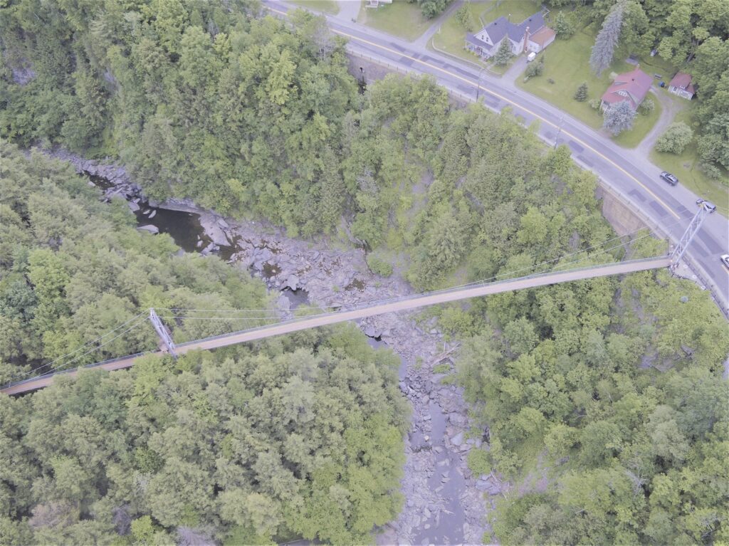 Vue aérienne d'une passerelle suspendue enjambant un ravin boisé avec un ruisseau rocheux en contrebas, reliant deux tronçons de route, avec des maisons et des voitures visibles en arrière-plan.