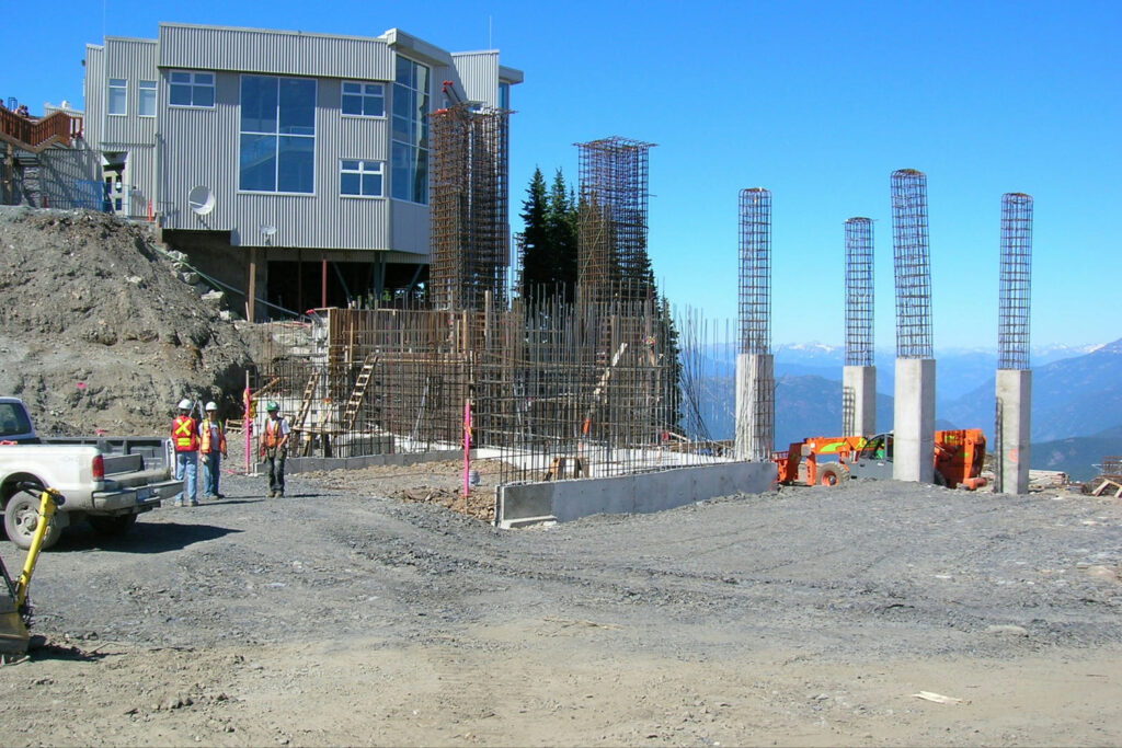 Construction site with workers in safety gear near partially built structures, including steel rebar columns and a large steel frame. A multi-story building and mountainous landscape are in the background.