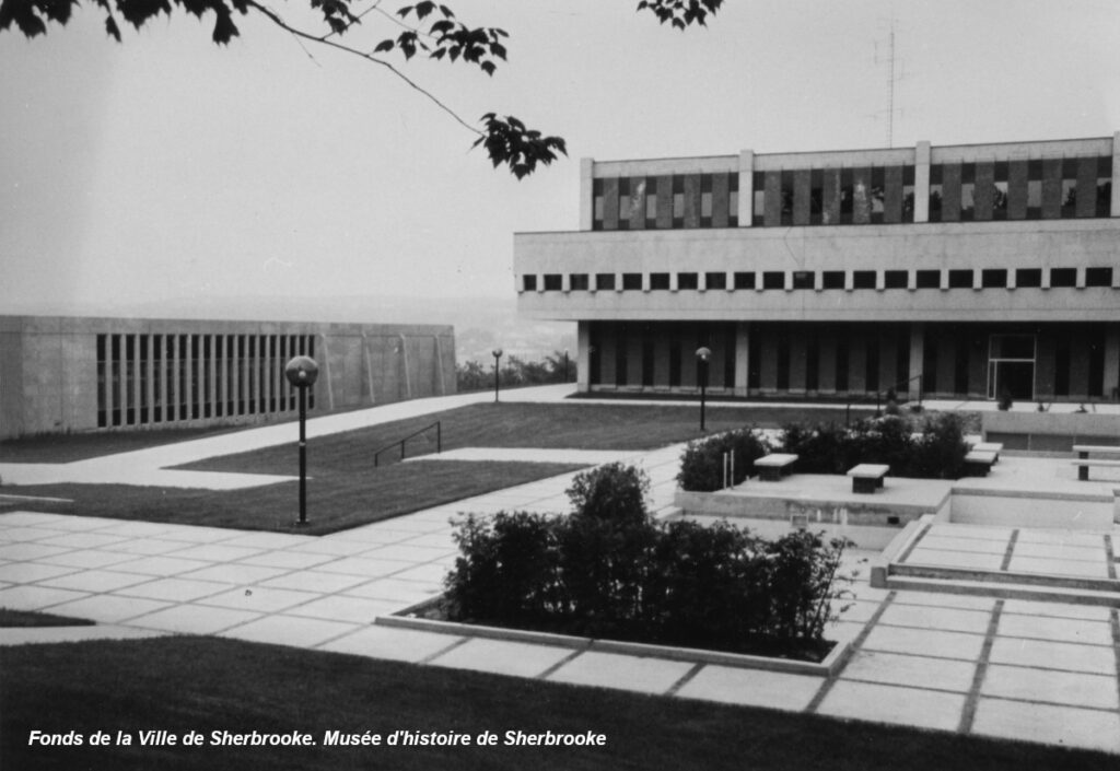 Photo en noir et blanc d'un bâtiment moderniste doté de fenêtres rectangulaires, entouré d'un espace gazonné et de passerelles en béton.