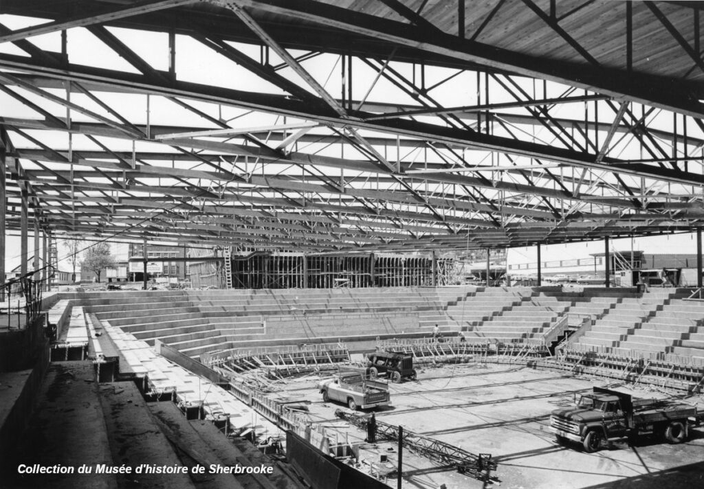 Photo en noir et blanc d'un stade couvert en construction avec des poutres en acier visibles et des sièges partiellement installés.