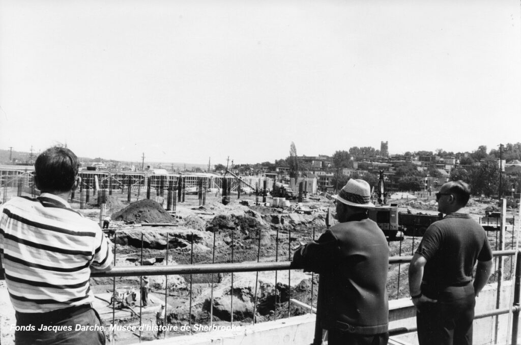 Three men observe a large construction site with multiple foundations in progress under a clear sky.