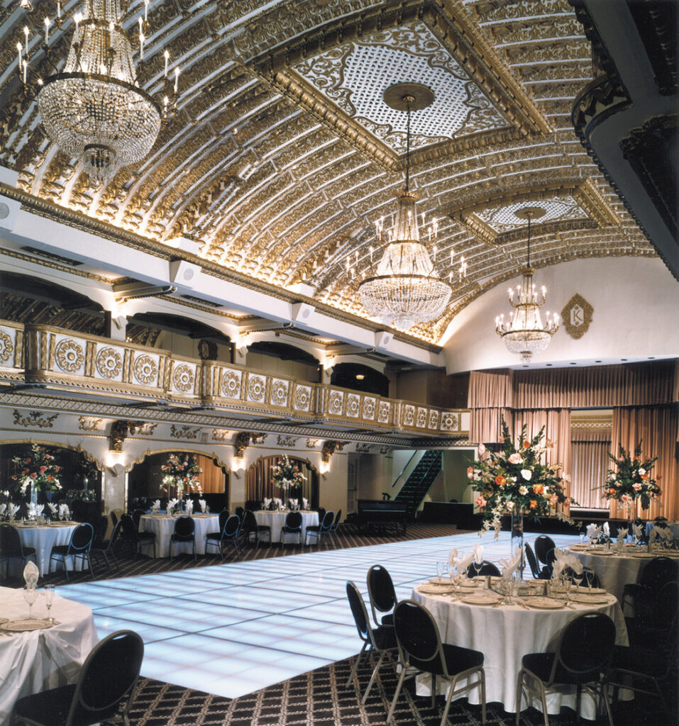 Une grande salle de bal avec un plafond doré orné, des lustres, des tables à manger rondes et un sol en damier. L'espace est aménagé pour un événement formel avec des centres de table floraux sur les tables.