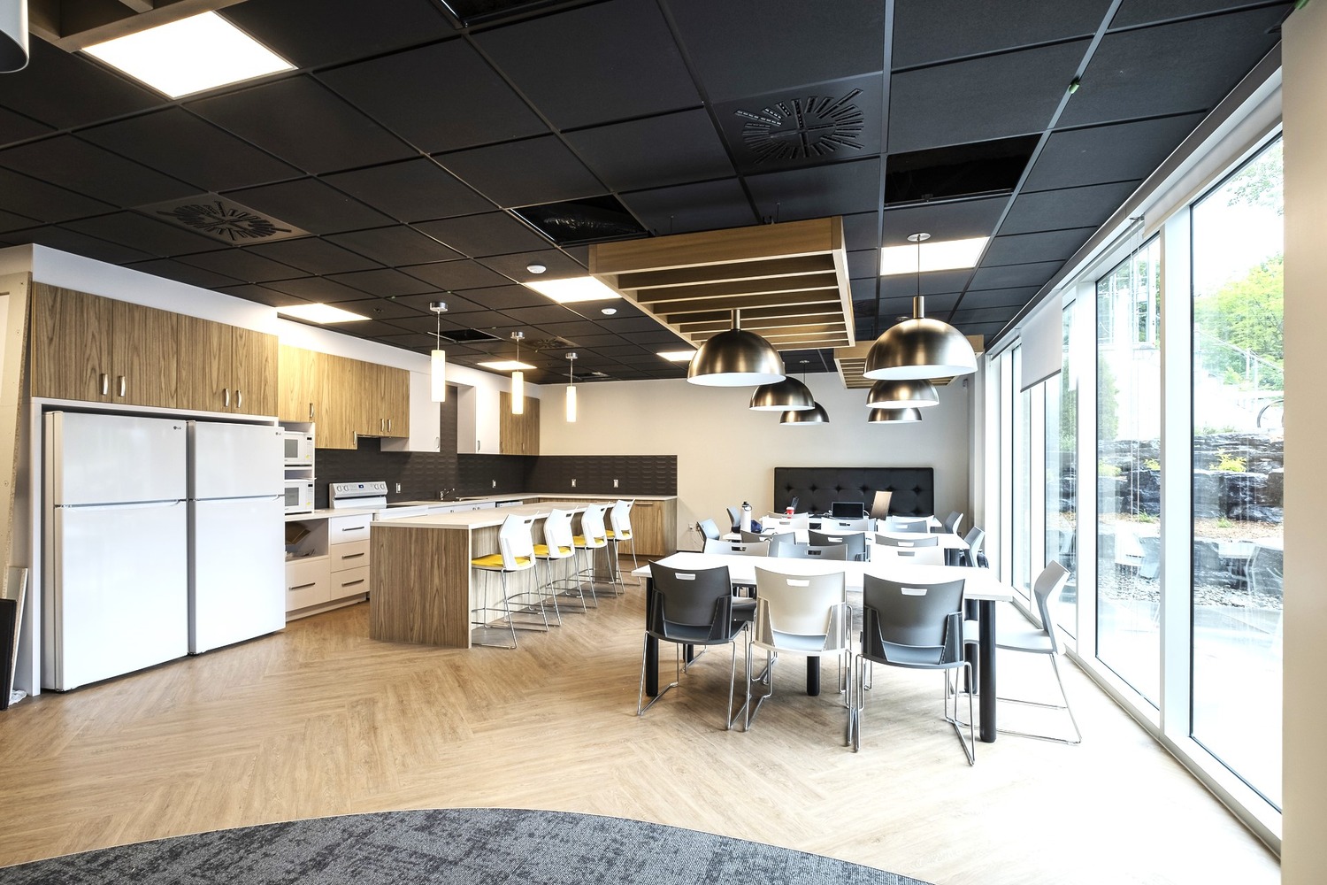 A modern communal kitchen and dining area with wooden floors, white and wooden cabinets, a large dining table with grey and white chairs, and pendant lighting. Large windows allow natural light to enter.