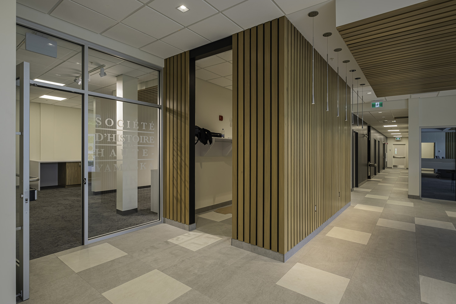 A modern office hallway with wooden panel accents and a glass door labeled "Société d'Histoire." The floor is tiled, and the ceiling features recessed lighting. The hallway leads to additional rooms.