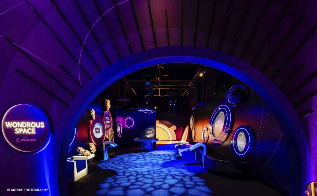 An illuminated exhibition space titled "Wondrous Space," displaying various circular informational panels and artifacts, with a futuristic and vibrant lighting design.