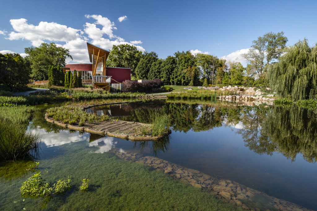 Un bâtiment moderne avec un haut toit triangulaire se dresse à côté d'un étang calme avec une allée en bois, entouré d'une verdure luxuriante et d'arbres sous un ciel bleu vif avec des nuages épars.