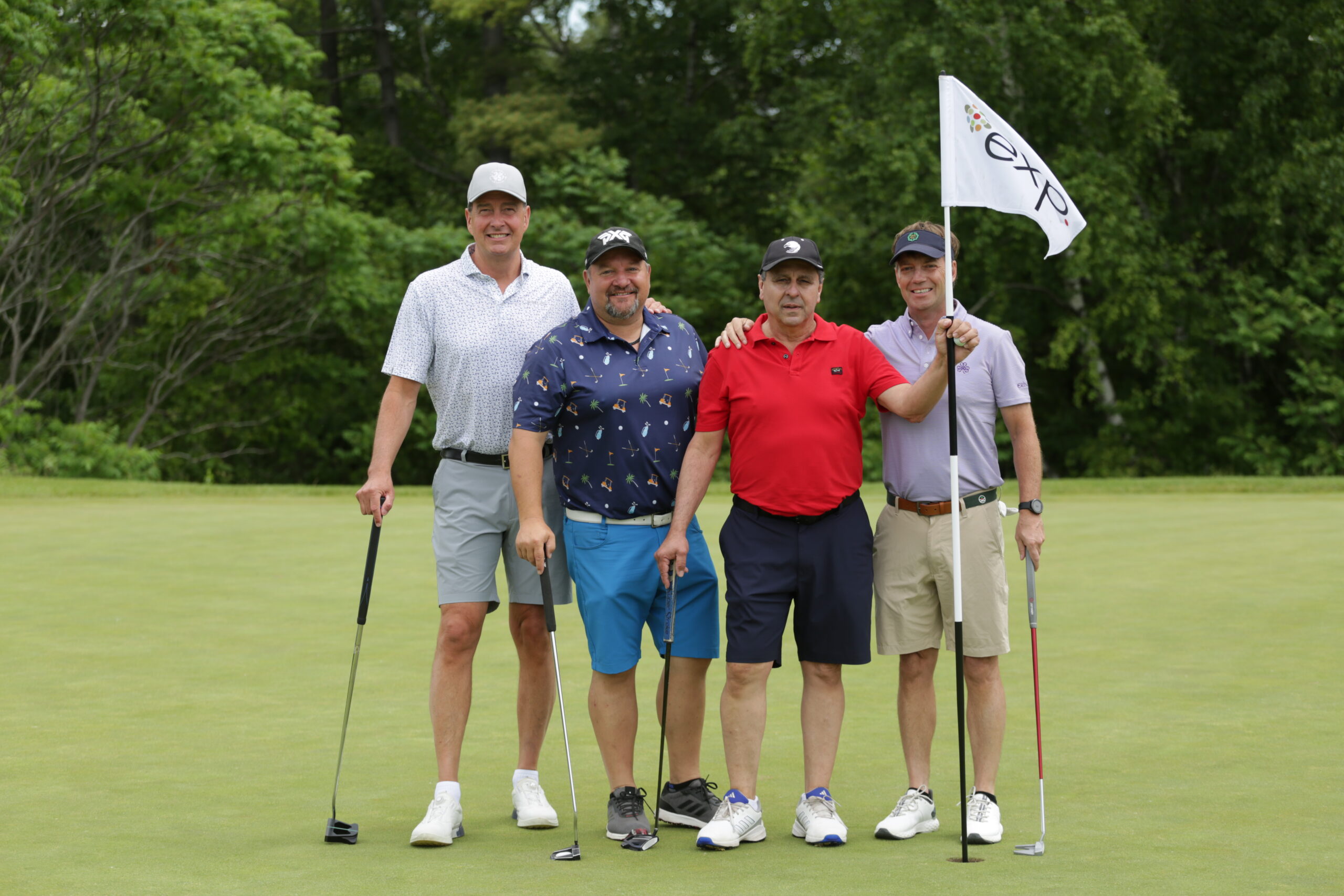 Four men pose on a golf course green, one holding a flagstick. They are dressed in golf attire with trees in the background.