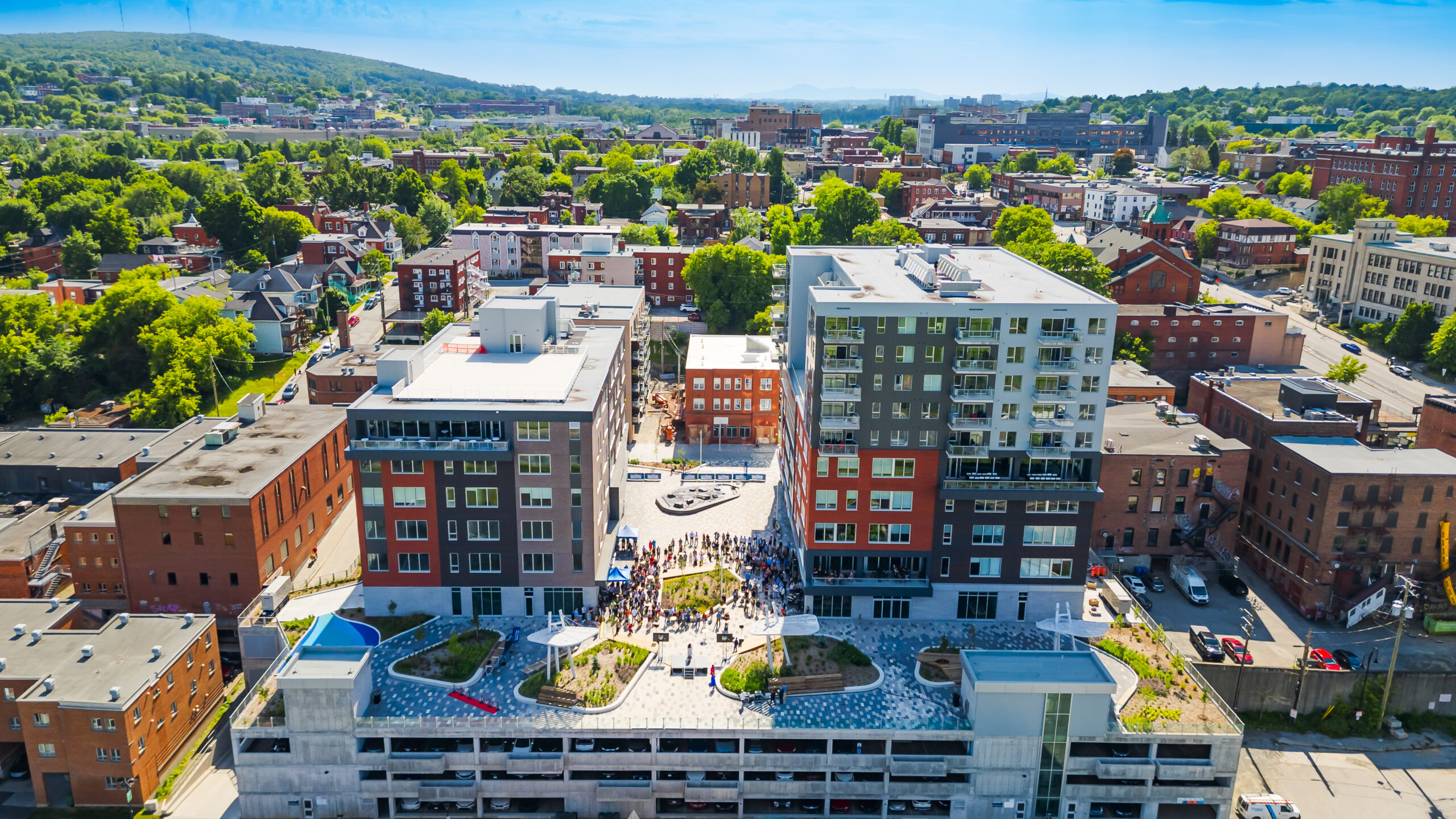 Vue aérienne d'un paysage urbain comprenant des bâtiments modernes avec des jardins sur les toits et une place centrale animée par les gens, entourée d'un mélange de verdure et de structures plus anciennes.