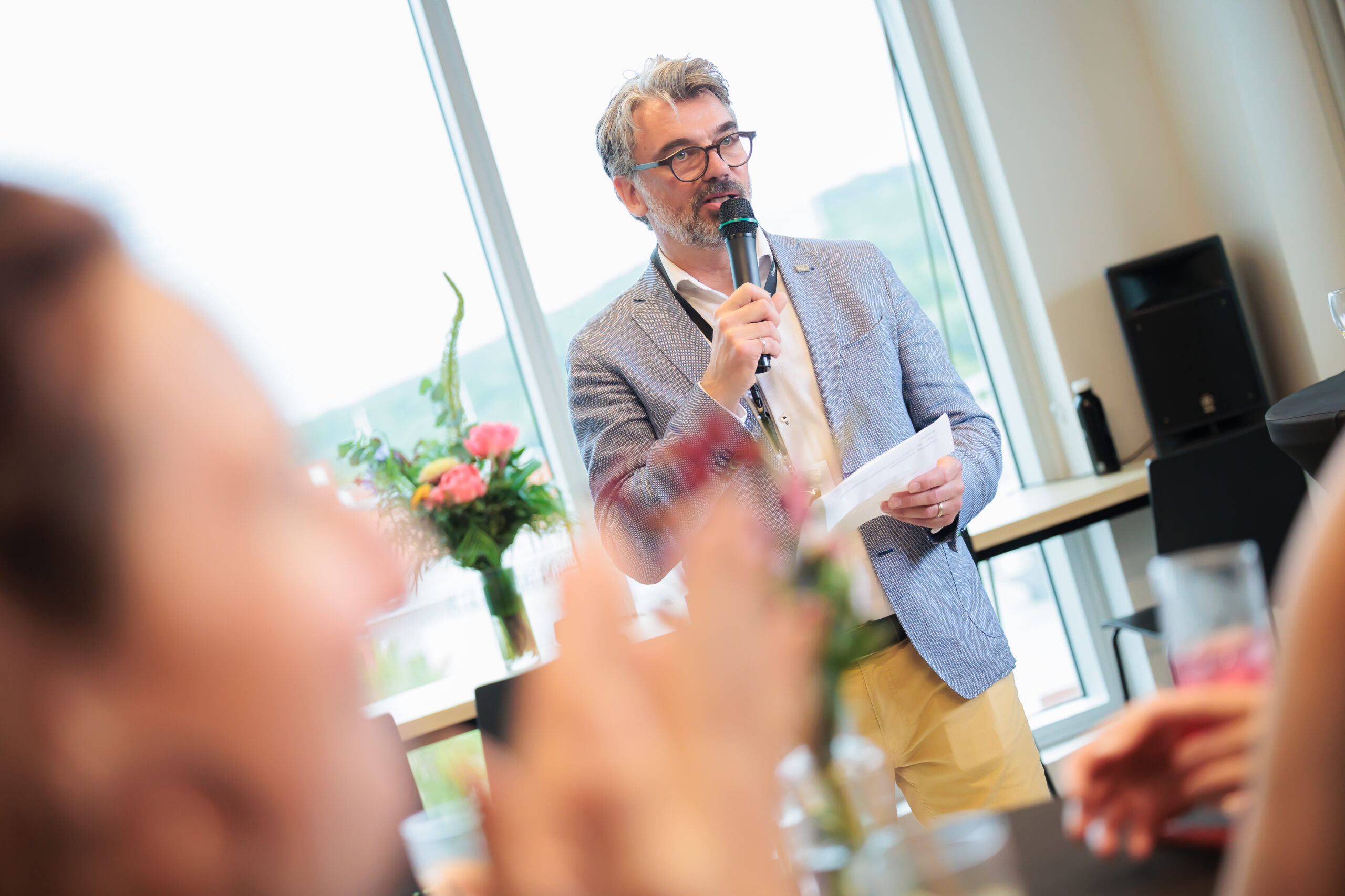 Un homme portant des lunettes et une barbe parle dans un microphone tout en tenant des papiers. Il se tient devant une grande fenêtre avec un vase de fleurs à proximité. Les membres flous du public sont visibles au premier plan.