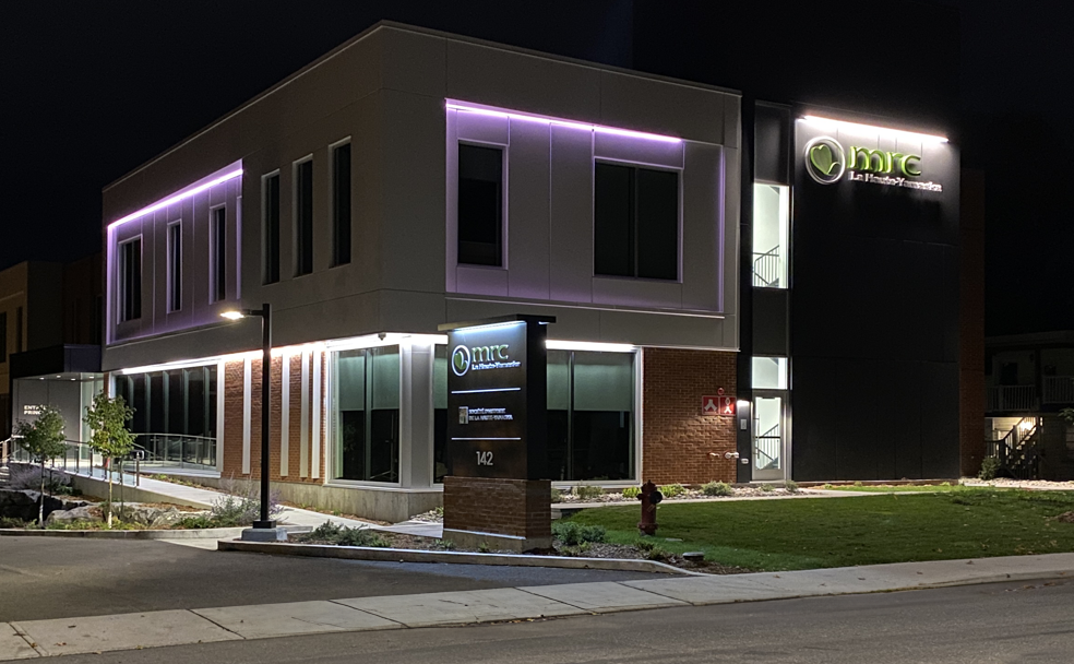 A modern two-story building with exterior lighting, labeled "MNC" Medical Center. The sign and building feature a green logo and the address 142 is visible on the sign in front.