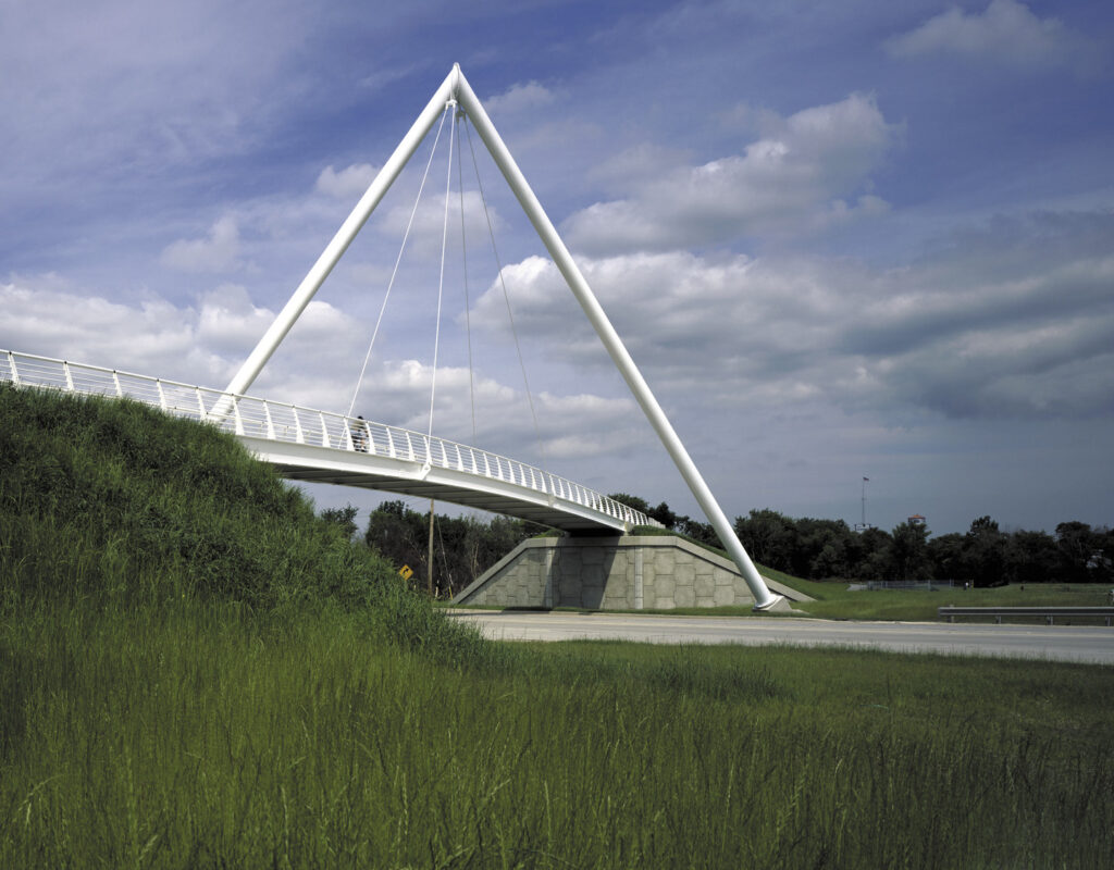 A white cable-stayed bridge with a triangular support spans over a green grassy area on a cloudy day.