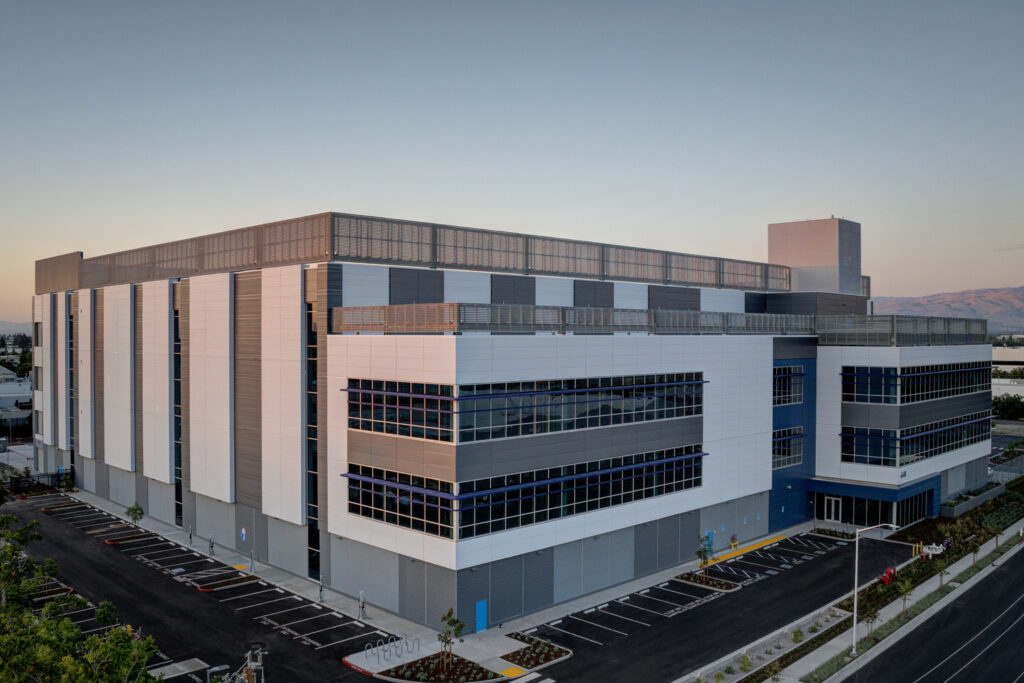 A modern, multi-story, rectangular building with numerous windows is shown. The building features a mix of white, gray, and blue exteriors and is situated in a parking lot with empty spaces.