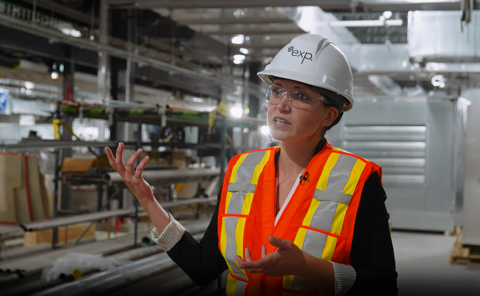 Une personne portant un casque blanc, des lunettes de sécurité et un gilet réfléchissant orange fait des gestes avec ses mains tout en parlant dans un environnement industriel.