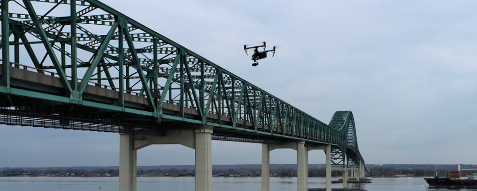 A drone is flying near a long, green steel bridge spanning over a body of water on a cloudy day.