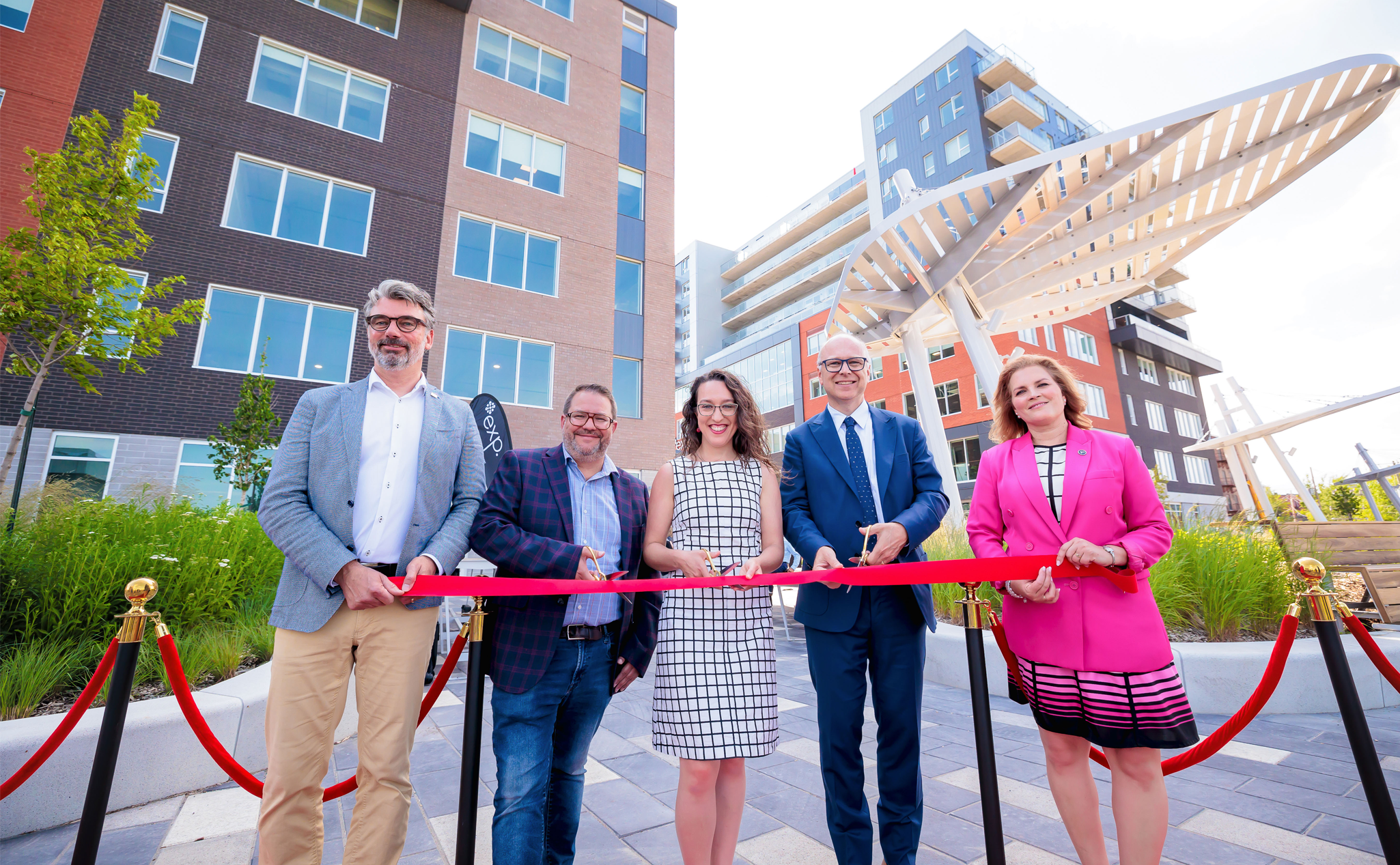 Five people stand in front of buildings, holding large scissors to cut a red ribbon for a ceremony. The setting appears to be outdoors under a sunny sky.