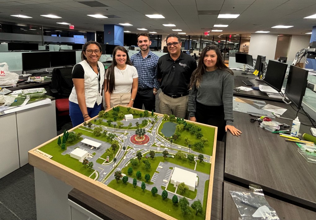 Five people stand around a detailed architectural model of a proposed urban development, featuring roads, buildings, and landscape greenery, on a table in an office setting.