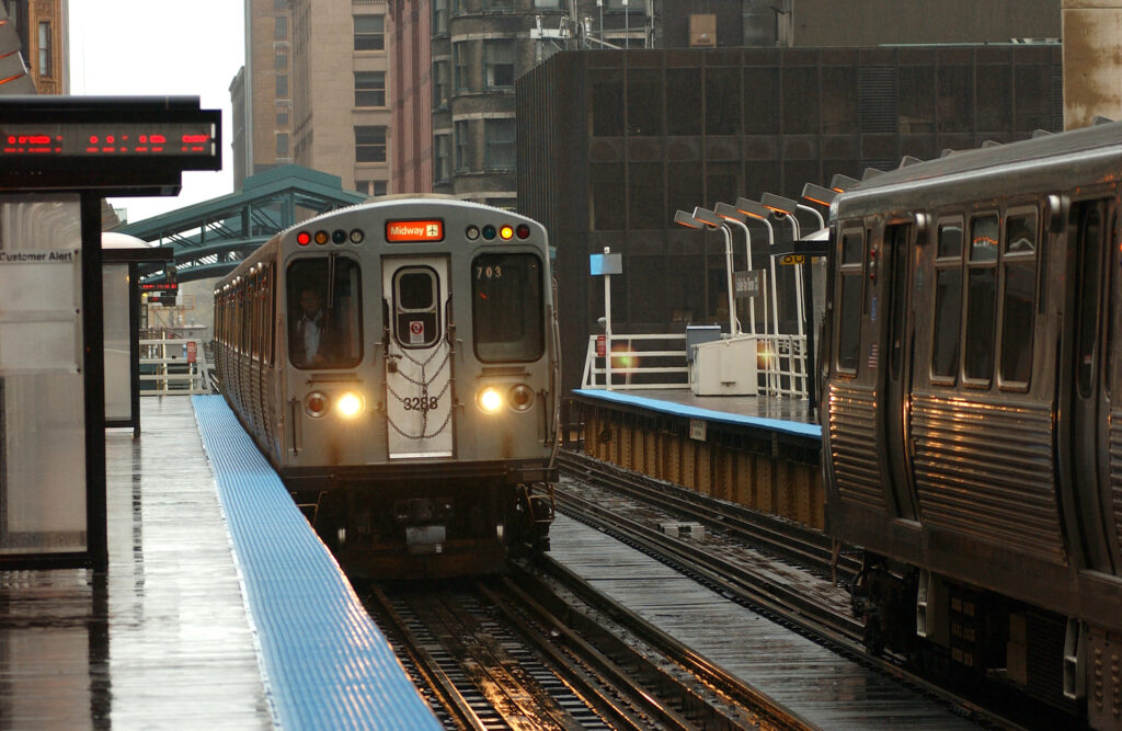 Deux trains dans une gare « L » de Chicago ; l'un arrivant par temps pluvieux tandis que l'autre est à l'arrêt. La scène comprend de grands immeubles en arrière-plan.