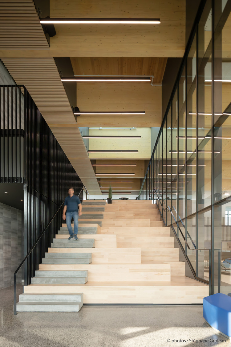 Une personne descend de larges escaliers en bois modernes avec des rampes en métal dans un intérieur spacieux et bien éclairé doté de grandes fenêtres et d'un haut plafond avec des poutres en bois.