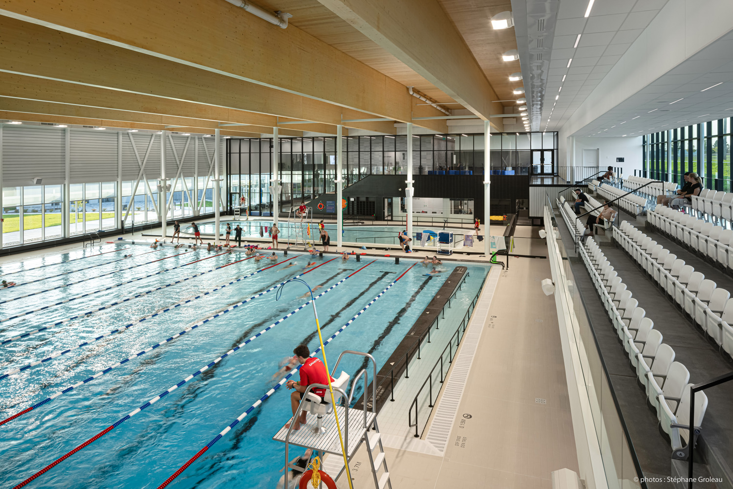 Piscine couverte avec plusieurs couloirs, nageurs et sauveteur en service. Les sièges des spectateurs se trouvent du côté droit. De hautes fenêtres et un éclairage lumineux remplissent l'espace.