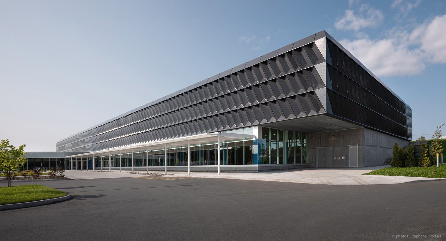 Un bâtiment moderne avec une façade géométrique unique, de grandes fenêtres et un surplomb couvrant la zone d'entrée. Le ciel est clair et il y a de petits espaces paysagers autour de la structure.