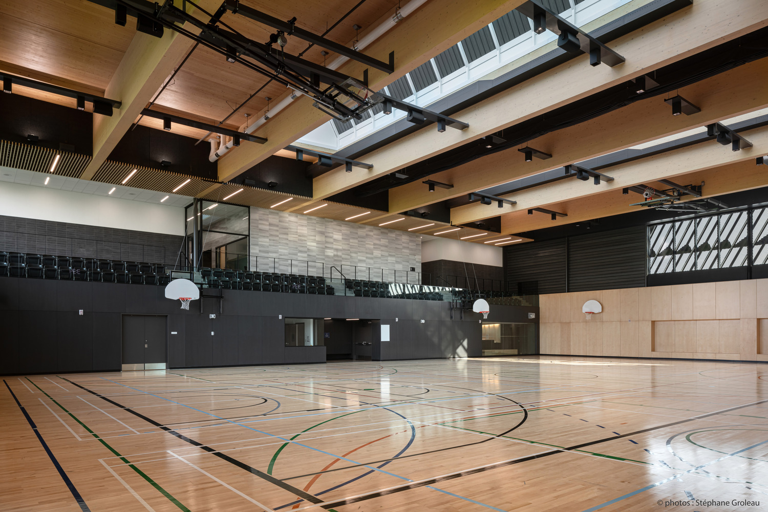 Un terrain de basket intérieur moderne avec un parquet, plusieurs paniers de basket et des sièges pour les spectateurs. La cour bénéficie d'un éclairage naturel provenant de lucarnes et d'un haut plafond avec poutres apparentes.